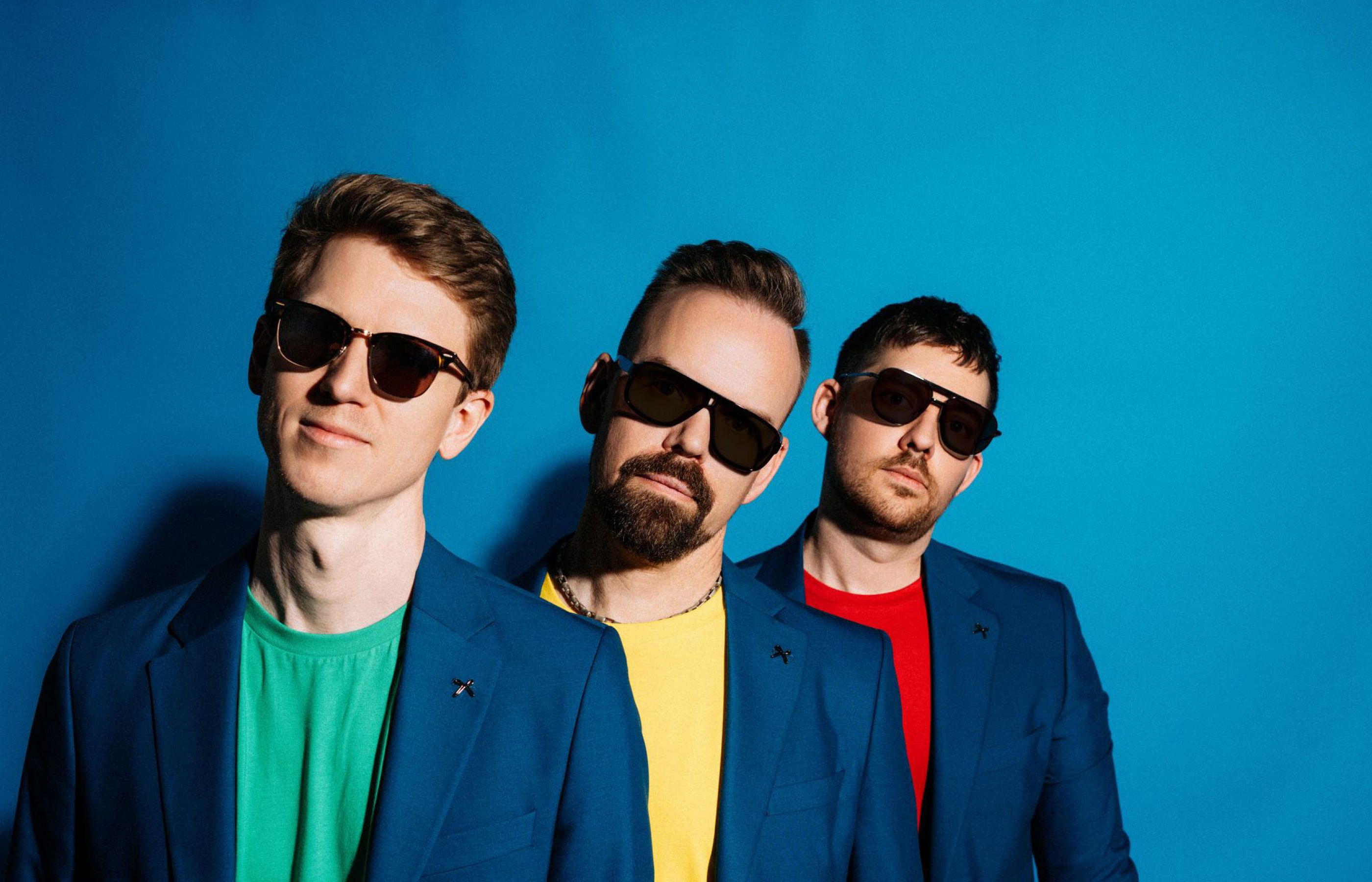 Die drei Zanadu Bandmitglieder mit bunten Tshirts vor blauem Hintergrund